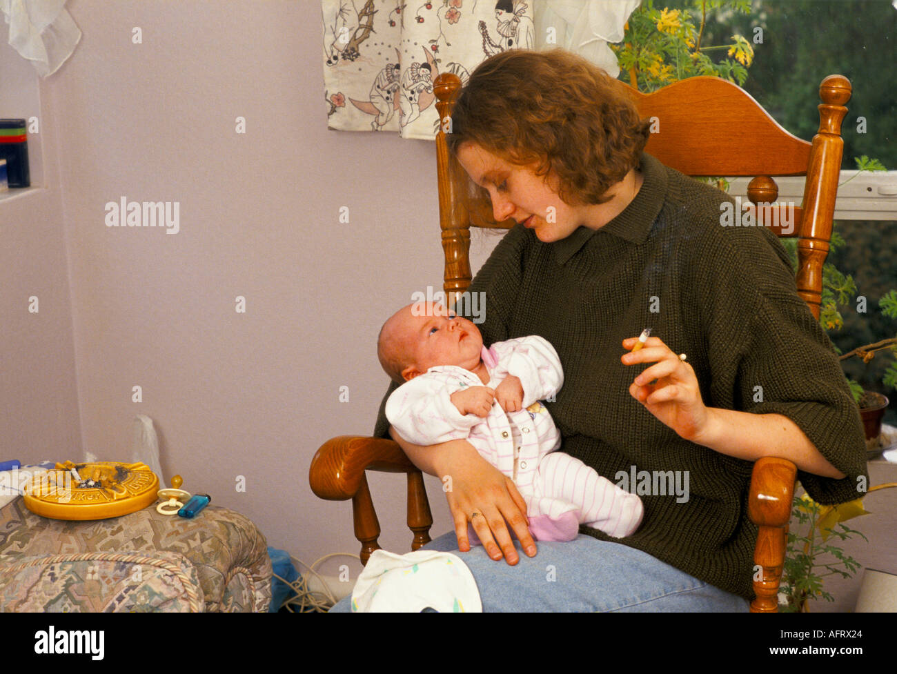 Young mother home with new four month old baby smoking a cigarette as