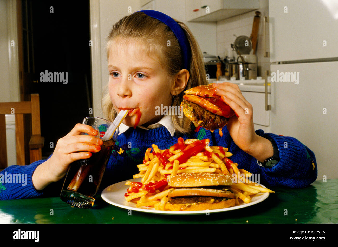 Young schoolgirl eating a huge hamburger and chips drinking coke at home in after school London. HOMER SYKES Stock Photo