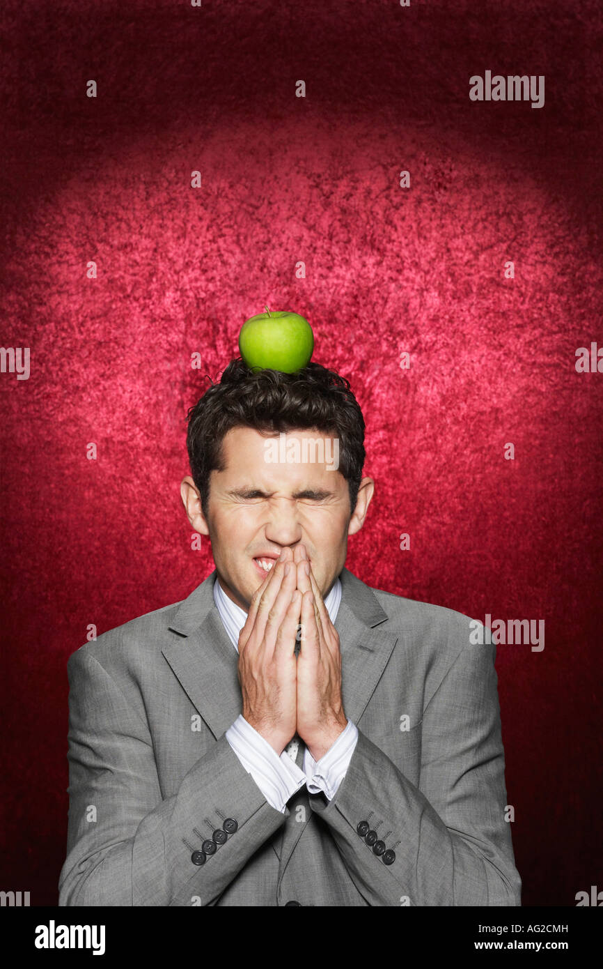 Man cringing with apple on head against red velvet background Stock Photo