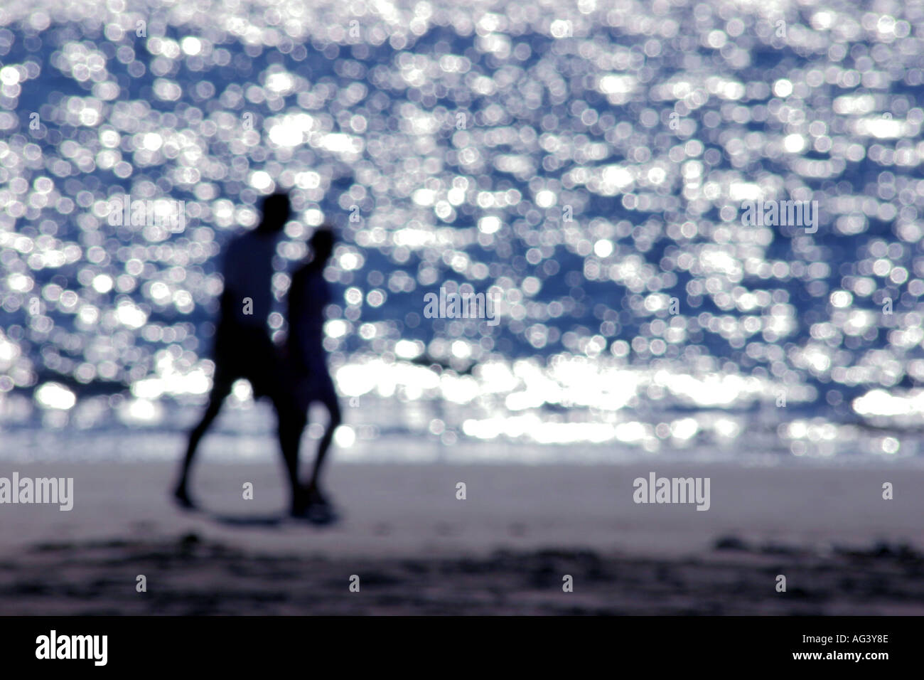 Couple walking along beach silhouetted against sea reflections blurred soft focus Stock Photo