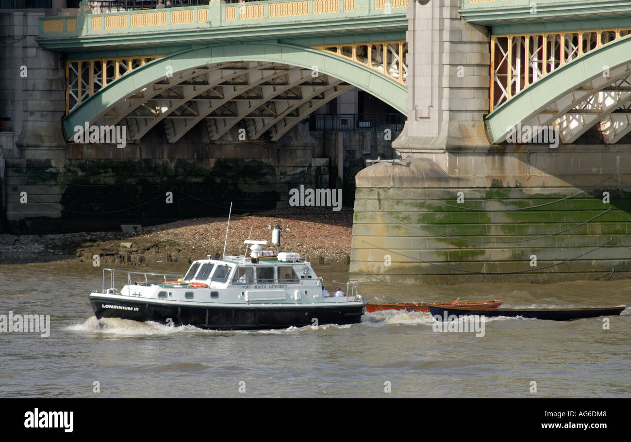 Two skiffs are towed up the River Thames under Southwark Bridge by the Port of Health Authority launch Londinium Stock Photo