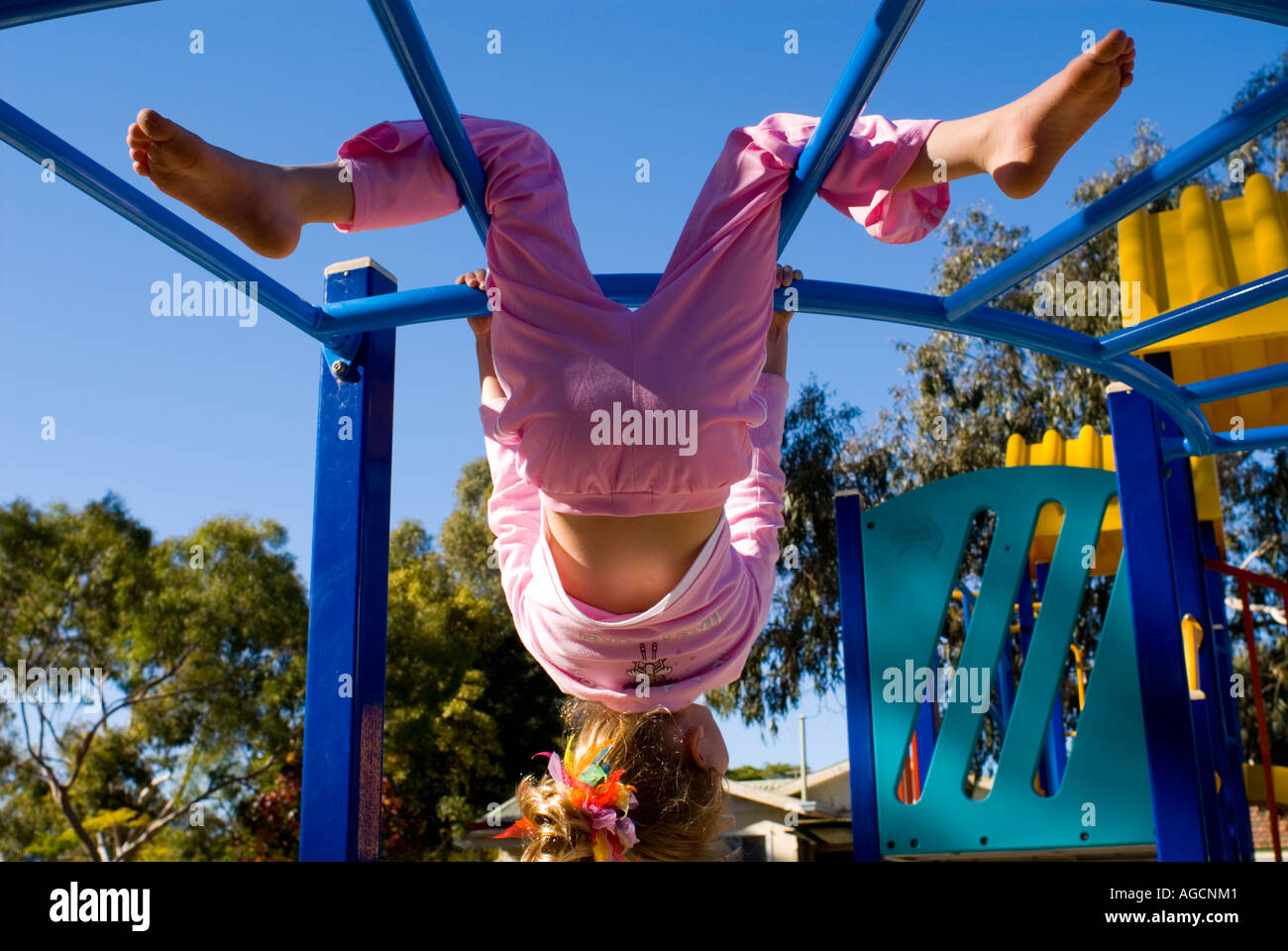 female child hanging upside down on playground climbing frame, against a deep blue sky Stock Photo