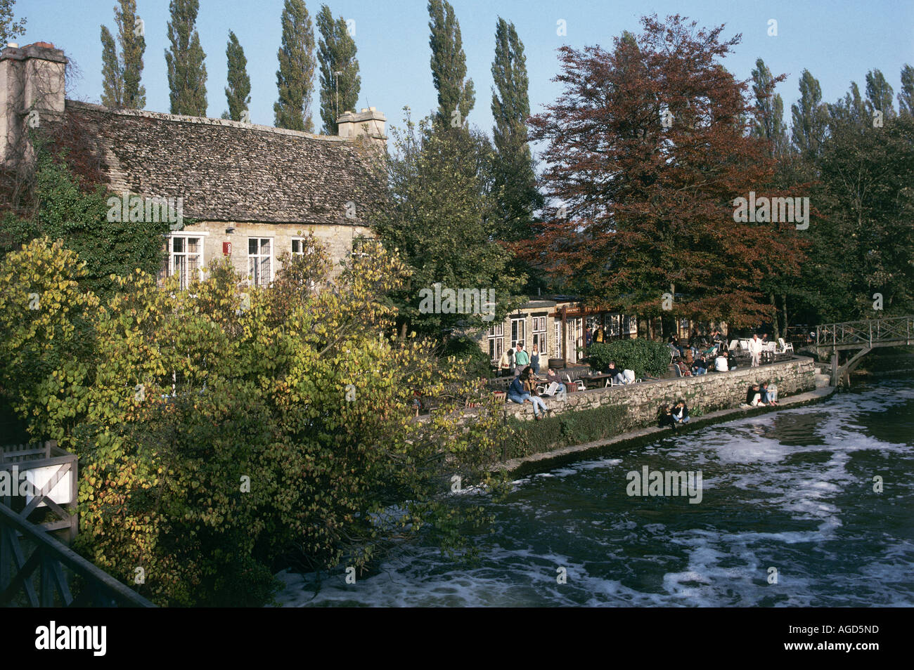 The stone built Trout public house on the banks of the Thames at Godstow stands upon the site of a traveller s hostel which was once used as a resting place for visitors to the nearby abbey People sit along the river bank White foam on the water Stock Photo