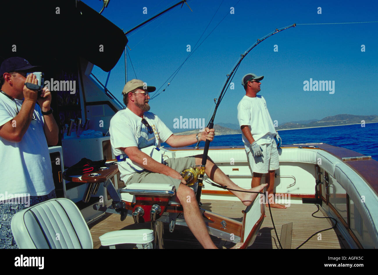Three men on the back of a fishing boat One man seated with a fishing pole  one filming with a video camera one man watching Stock Photo - Alamy