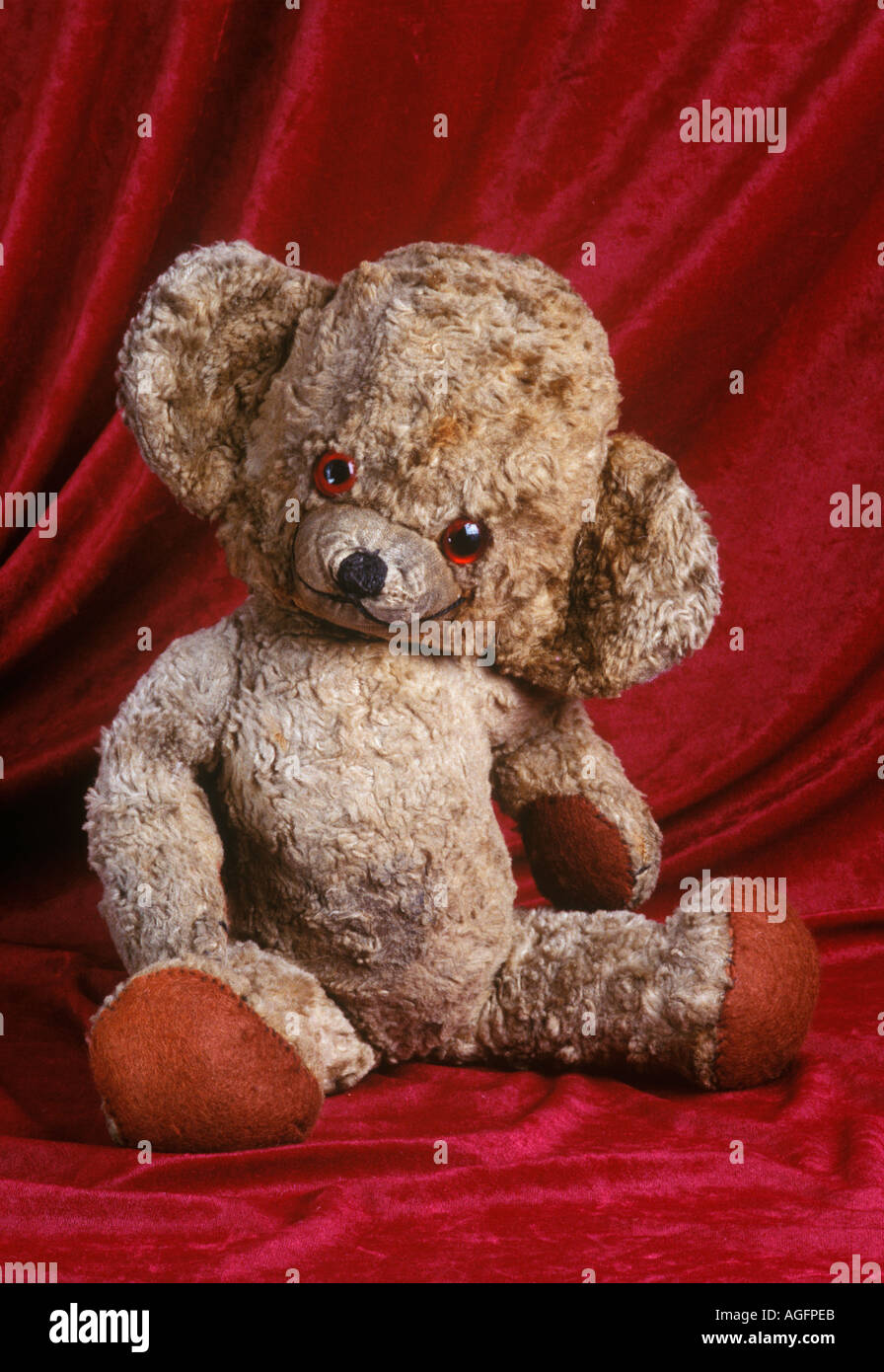Old scruffy well loved teddy bear sitting against red draped background Stock Photo