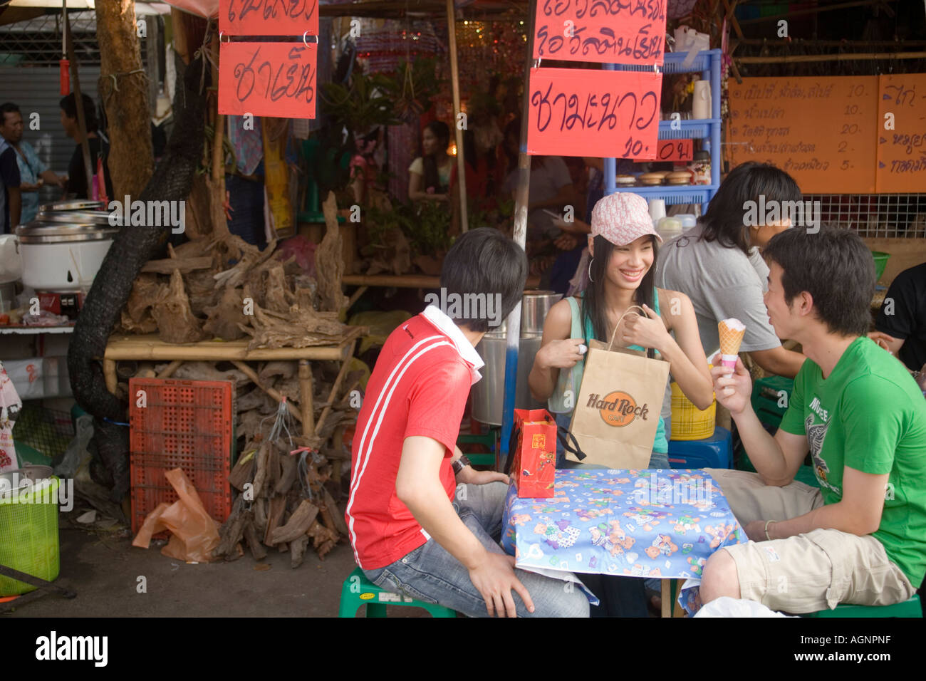 Three young people sitting at a table near a food stall one eating ice cream Suan Chatuchak Weekend Market Bangkok Thailand Stock Photo