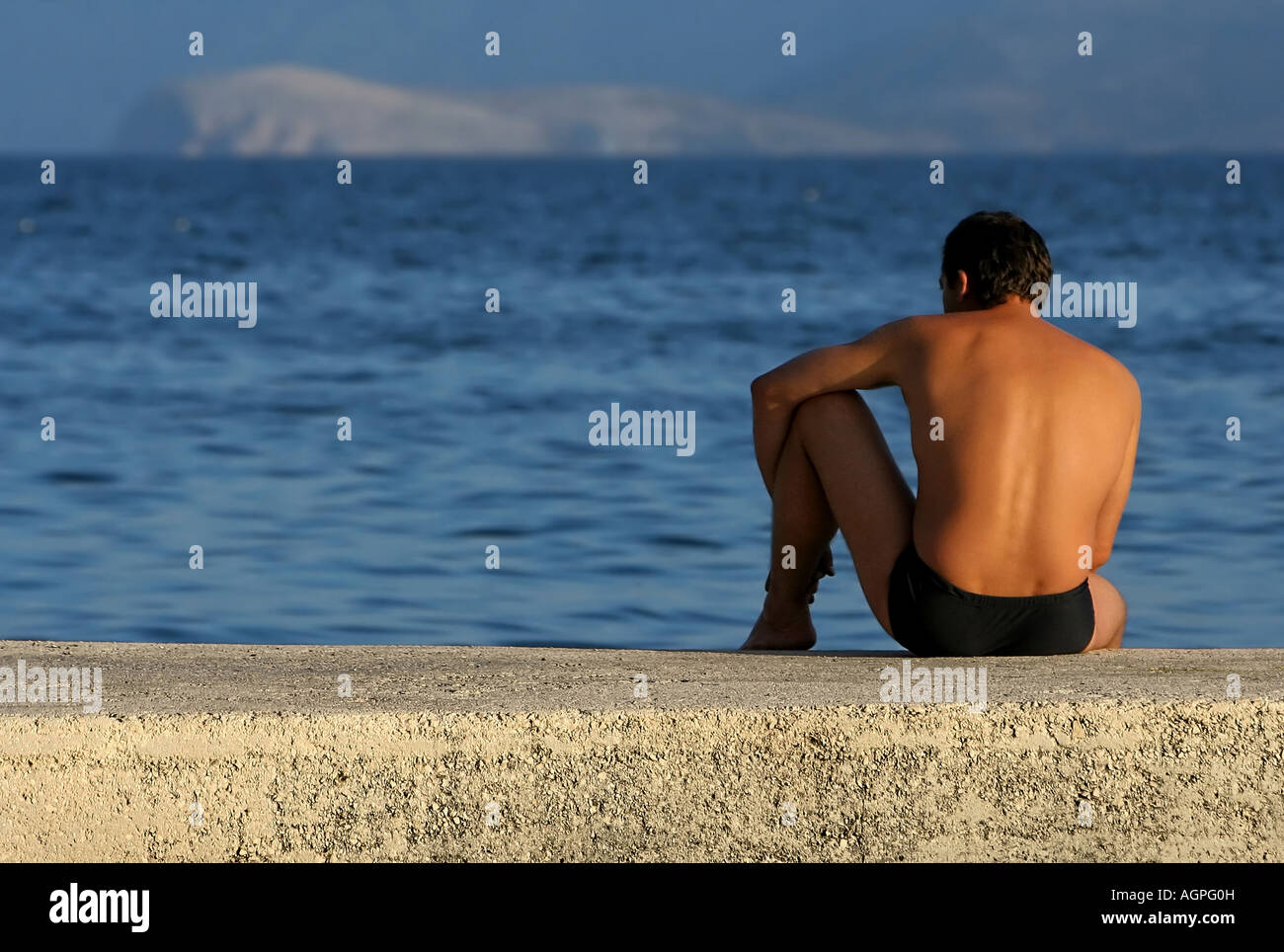 a man in bathing trunks sitting on a wall at the seashore and relaxing in the late afternoon sun Stock Photo