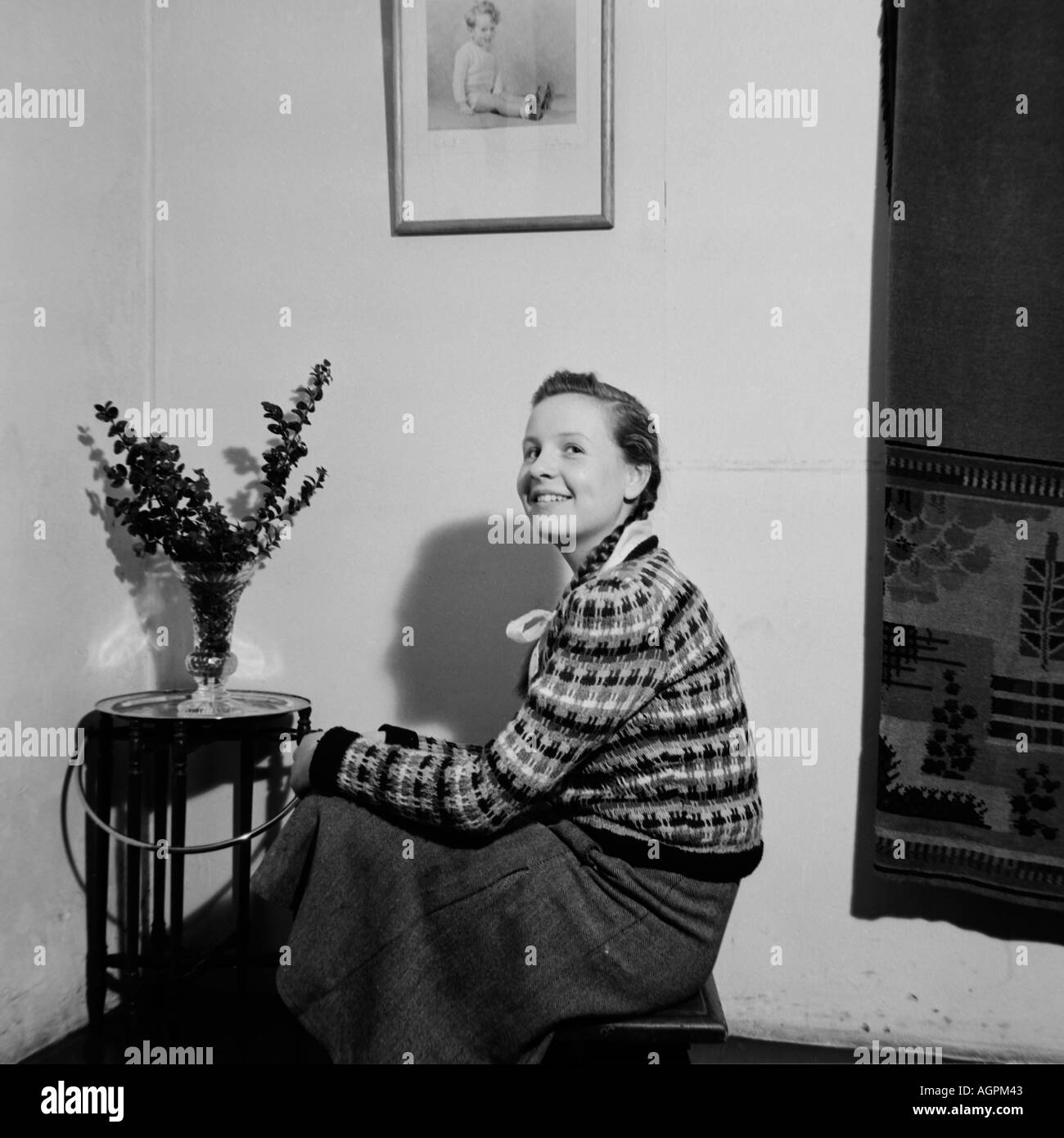 OLD VINTAGE FAMILY PHOTOGRAPH SNAP SHOT GIRL SITTING ON STOOL NEXT TO TABLE WITH VASE OF FLOWERS CIRCA 1950 Stock Photo