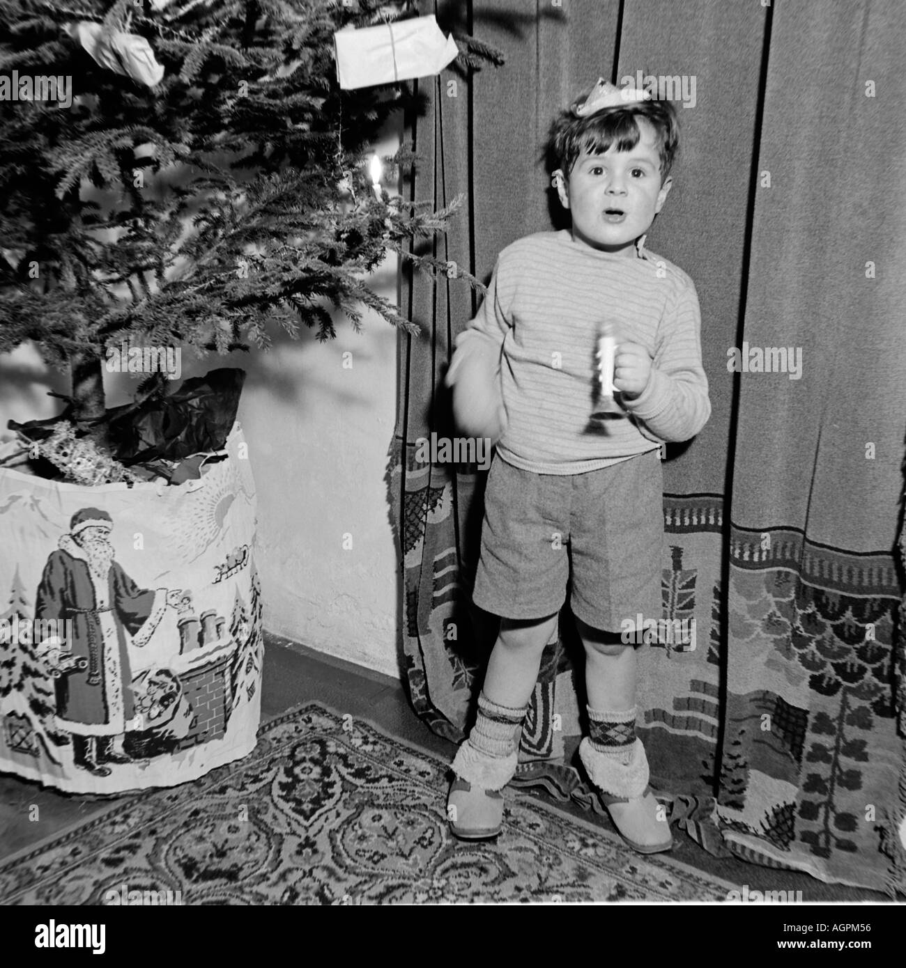 OLD VINTAGE FAMILY SNAPSHOT PHOTOGRAPH OF YOUNG BOY WEARING PARTY HAT PLAYING WITH TOY TRUMPET NEXT TO CHRISTMAS TREE Stock Photo