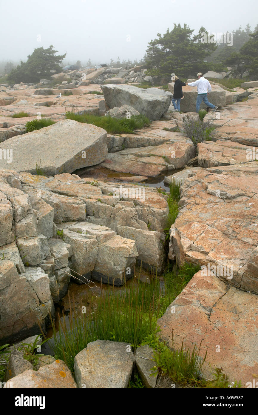 Caucasian 60 s couple step carefully over foggy stone crevices oceanside Schoodic Peninsula Acadia National Park Maine USA Stock Photo