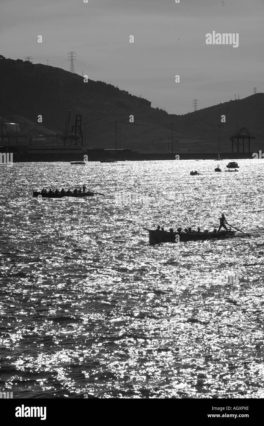 Traineras silhouetted by sun off Puerto Viejo de Algorta Basque Country Spain Hills in background Monochrome image Stock Photo