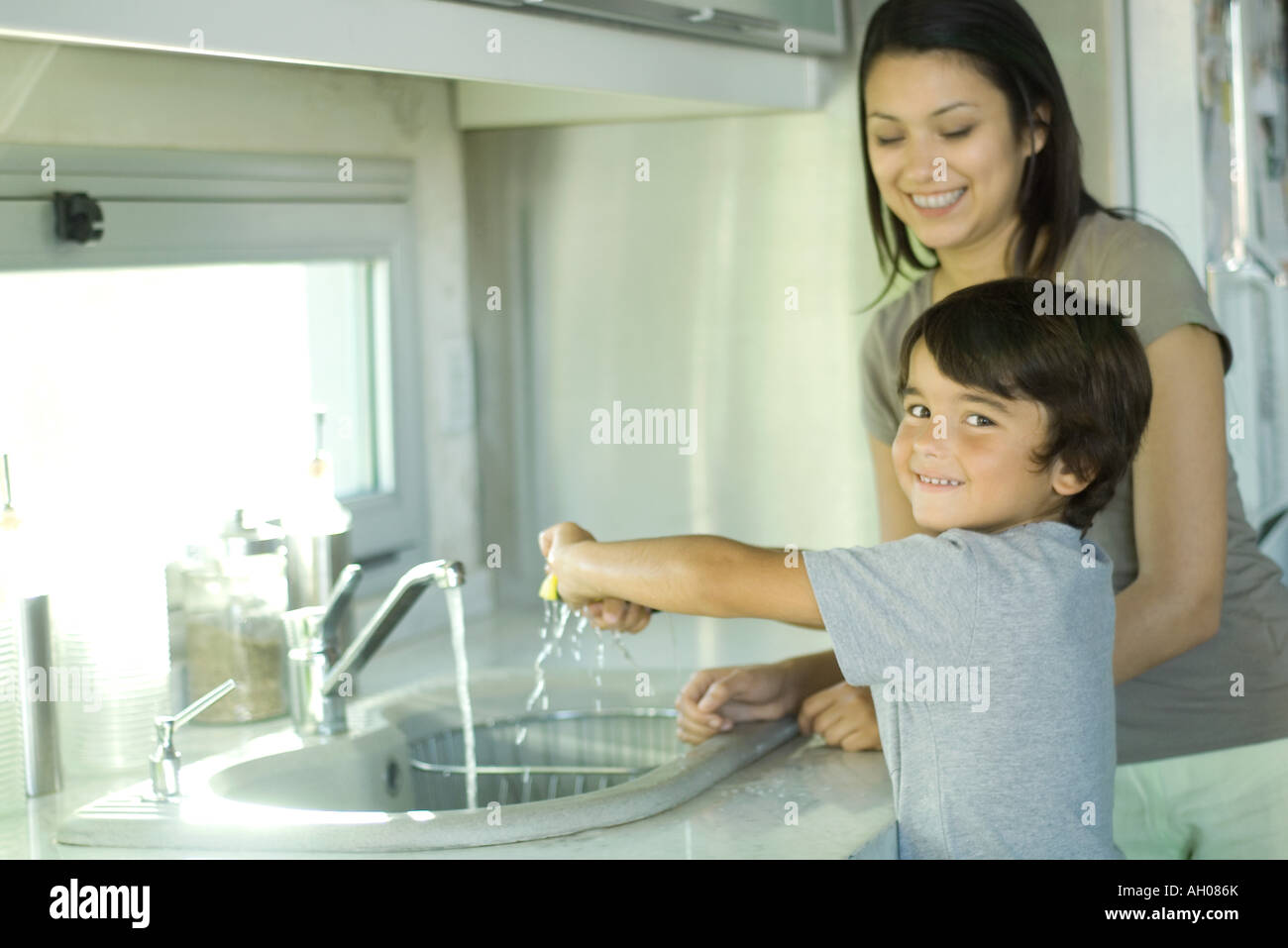 Boy Helping Mother Wash Dishes Looking Over Shoulder At Camera Sto