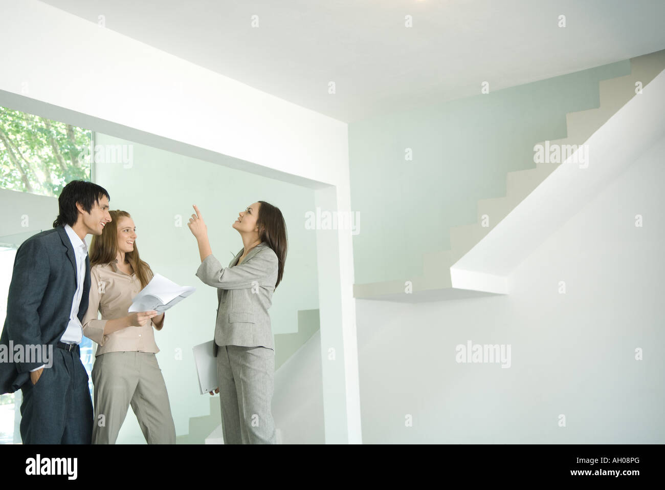 Female real estate agent showing house to young couple, pointing to ceiling Stock Photo