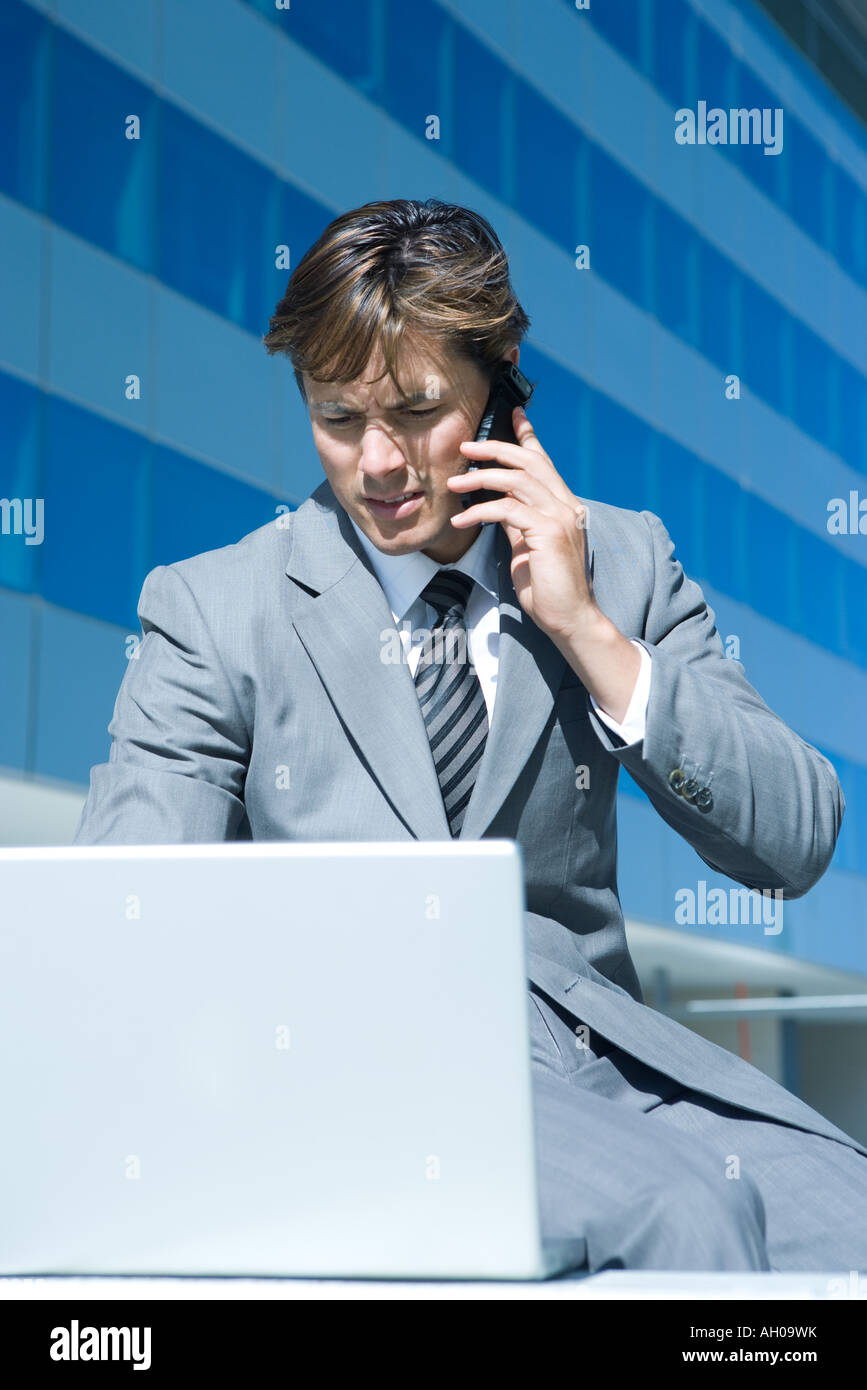 Young businessman sitting outdoors, using laptop computer, holding up cell phone Stock Photo