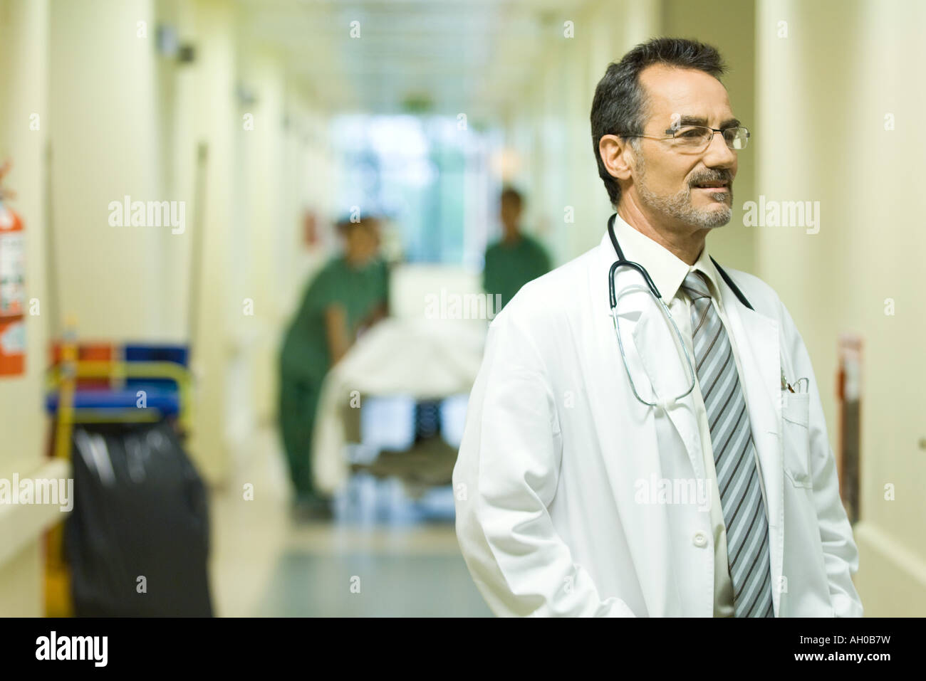 Male doctor looking away, smiling, hospital corridor in background ...