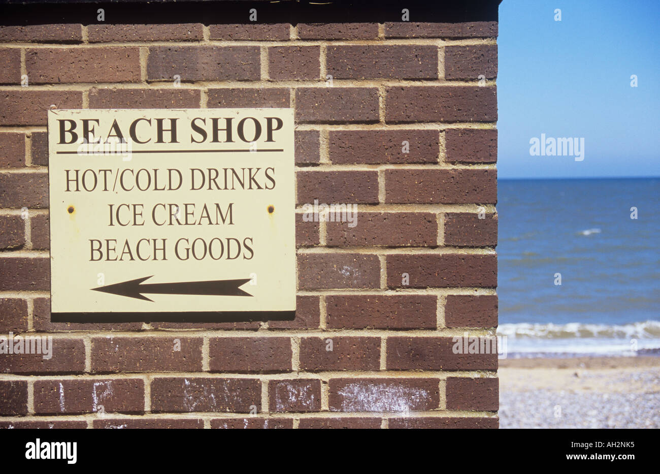 Rear of brick building with sign with arrow stating Beach Shop Hot cold drinks Ice cream Beach goods and with empty beach beyond Stock Photo