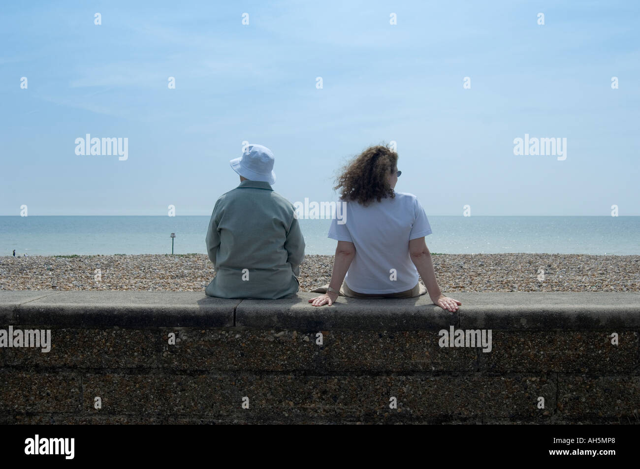 back view of two women sitting on beachfront wall looking out to sea in ...