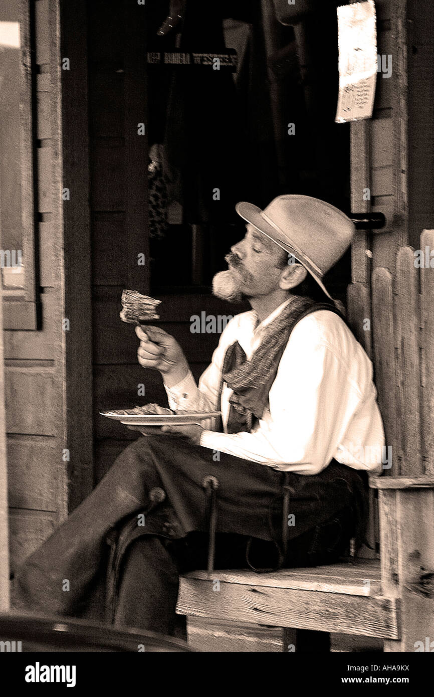 Cowboy Inspects Morsel While Eating Meal Outside Store On Bench In