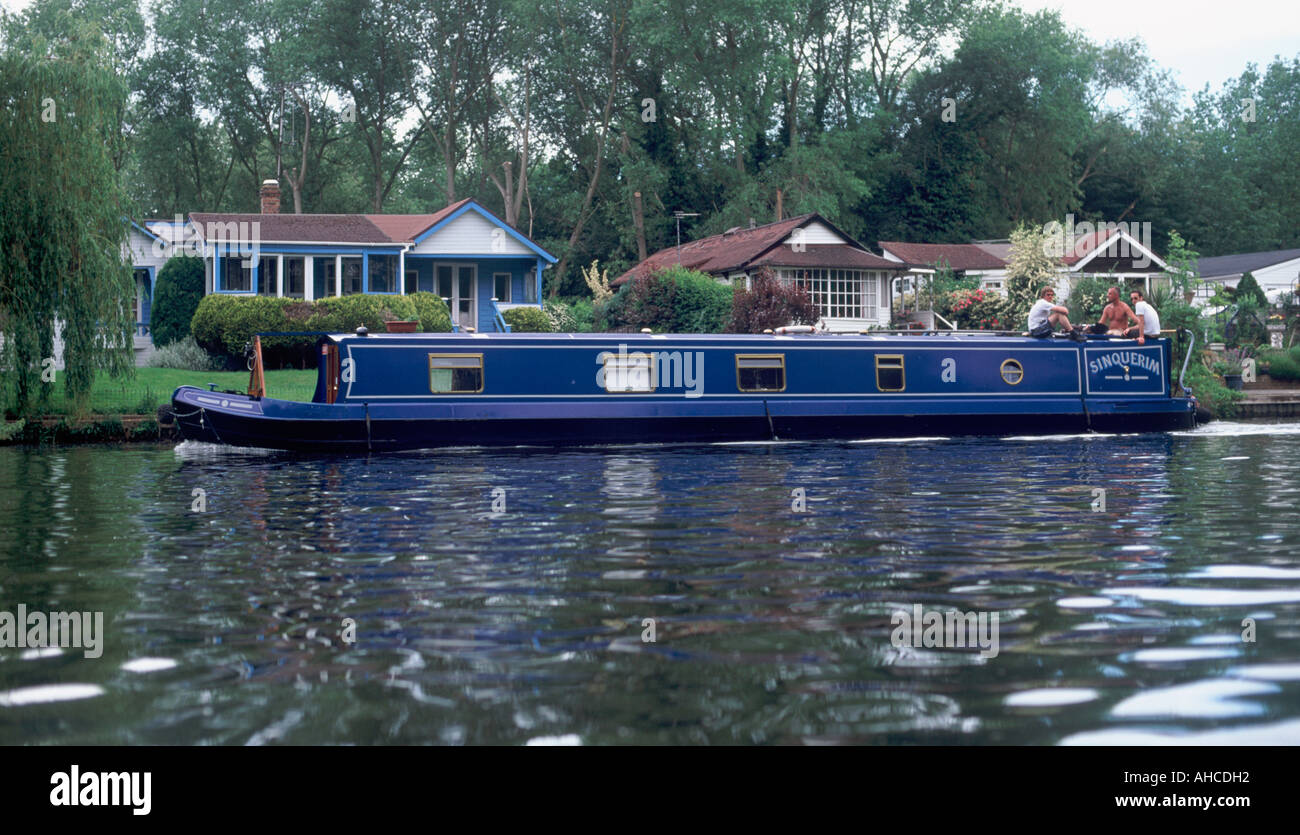 Traditional narrowboat on the River Thames passing riverside cottages, with three men at stern, Shepperton, Surrey, England Stock Photo