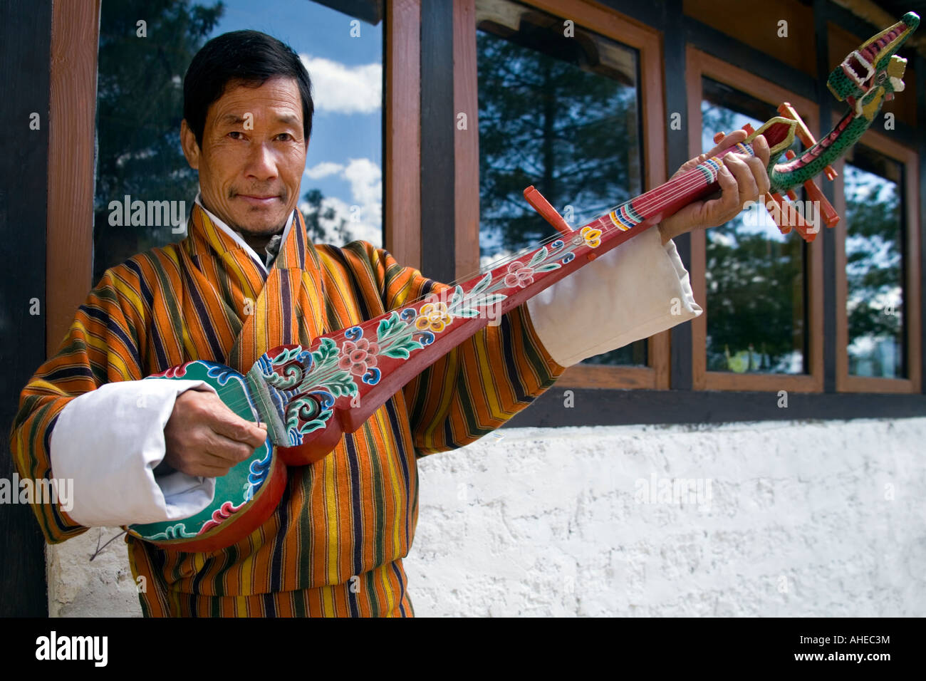 Portrait of a Bhutanese man in natiional dress wearing a warm coloured gho and playing a dramgyan lute at Taktshang teahouse Stock Photo