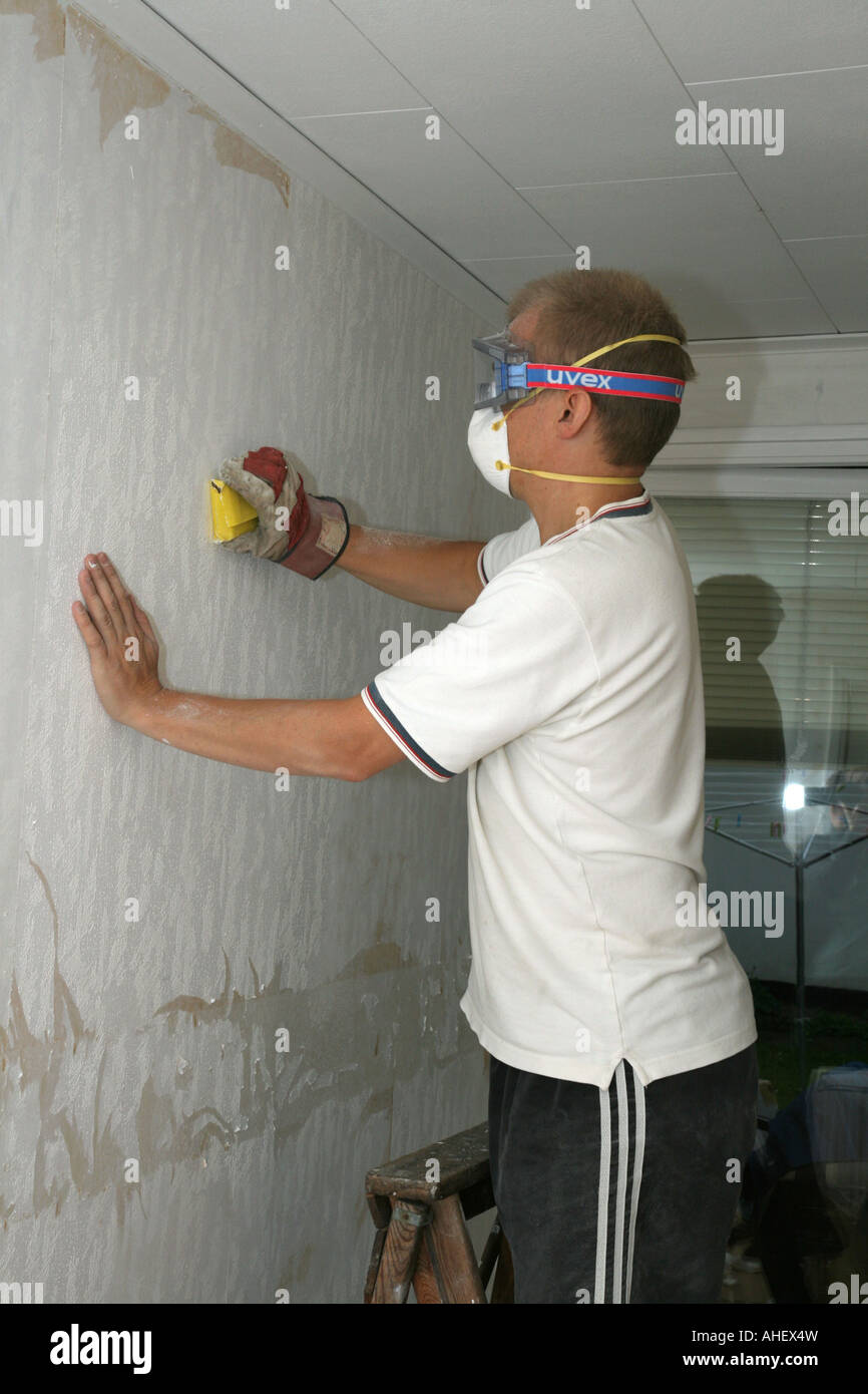 Man sandpapering a wall before putting up new wallpaper He is wearing