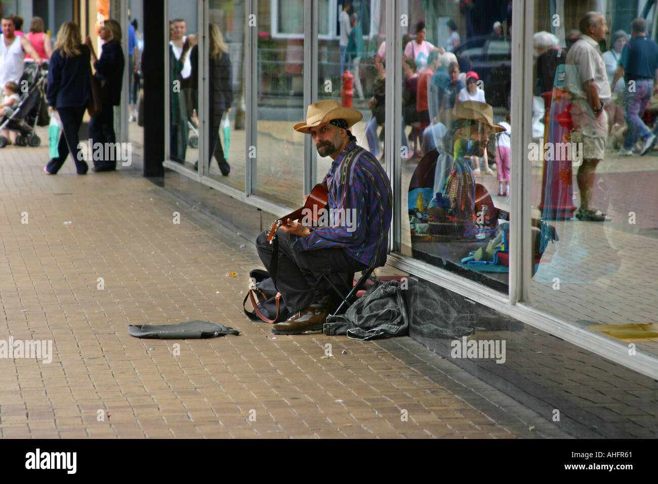 Man with hat and guitar squatting against shop window and busking for money Stock Photo