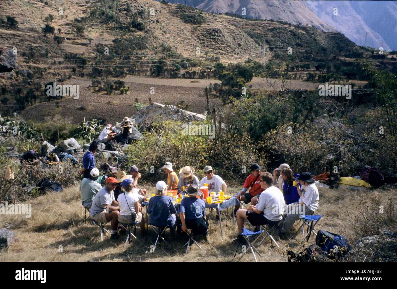 Group of tourists eating lunch at a folding table whilst guides and porters eat sitting on rocks behind. Inca Trail, Peru. Stock Photo