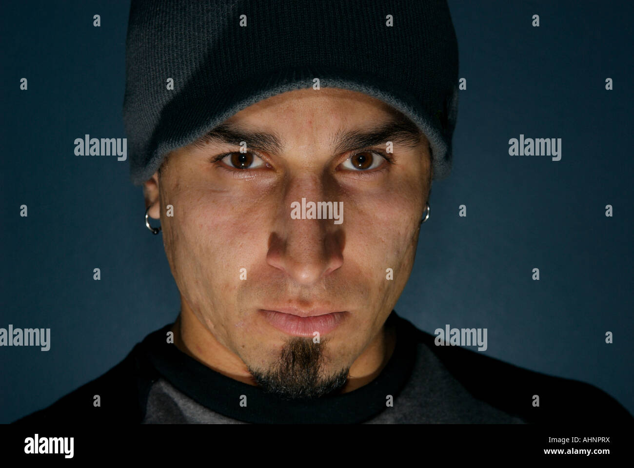A glaring Hispanic man with brown eyes and a goatee wearing earings and a beanie Stock Photo