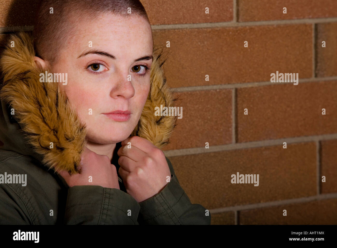 Teen girl with shaved head holding onto hood of coat and leaning on brick wall Stock Photo