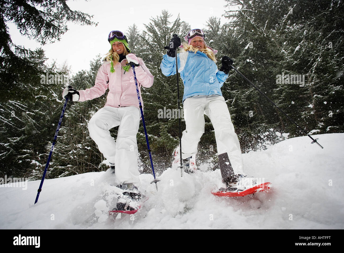 Low angle view of two young blonde women walking down small hill in forest using snow-shoes Stock Photo