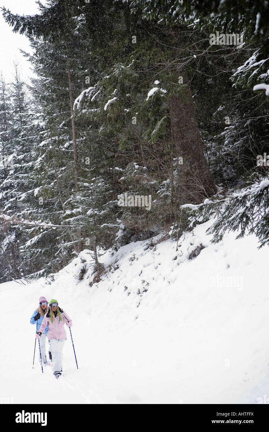 Two young blonde women walking down small hill in forest using snow-shoes Stock Photo