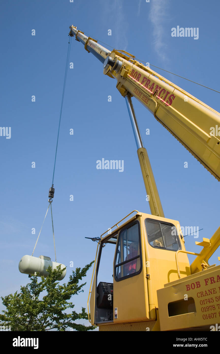 Calor gas tank being hoisted above trees by a yellow crane from a garden against blue sky. UK, Britain Stock Photo
