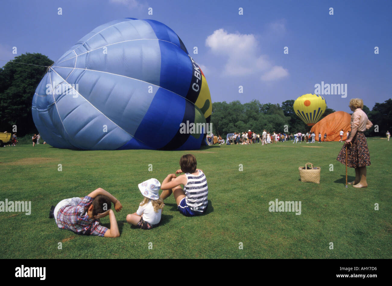 Age spectrum young children sit on grass & senior mature woman with walking stick watching hot air balloon launch on blue sky day Leeds Castle Kent UK Stock Photo