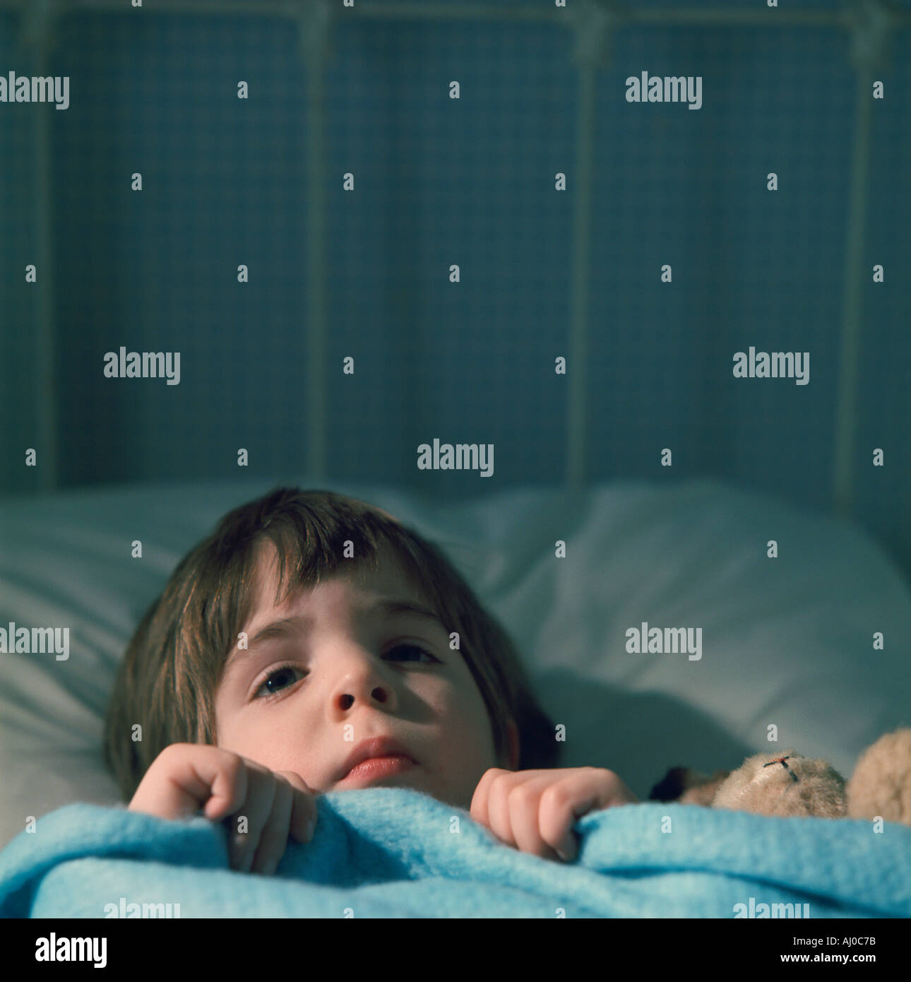 Young child lies in bed with a teddy bear and the covers pulled up to his chin Stock Photo