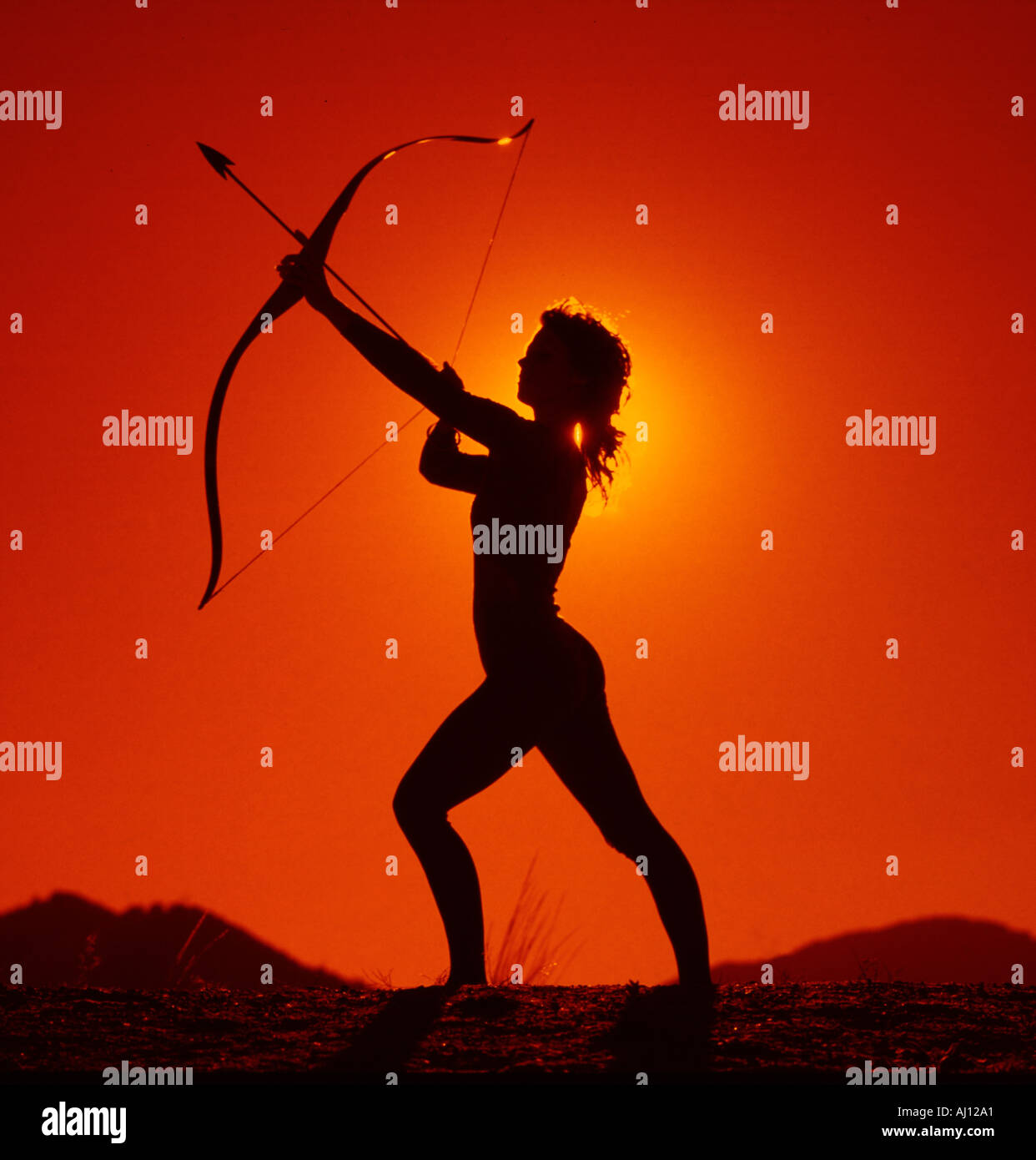Sunset silhouette of dramatic and agressive pose of young woman aiming a bow and arrow Stock Photo