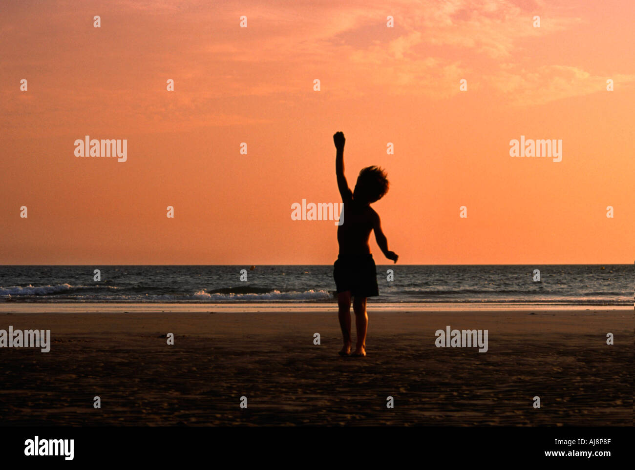 silhouette of young boy stretching up in the air while playing on the beach at sunset Stock Photo