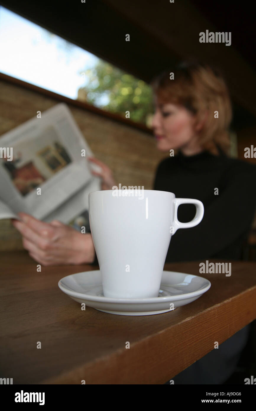 Coffee cup on table in cafe with woman sitting in background reading newspaper Stock Photo