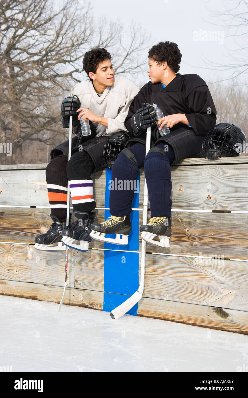 Two boys in ice hockey uniforms sitting on ice rink sidelines talking to eachother Stock Photo