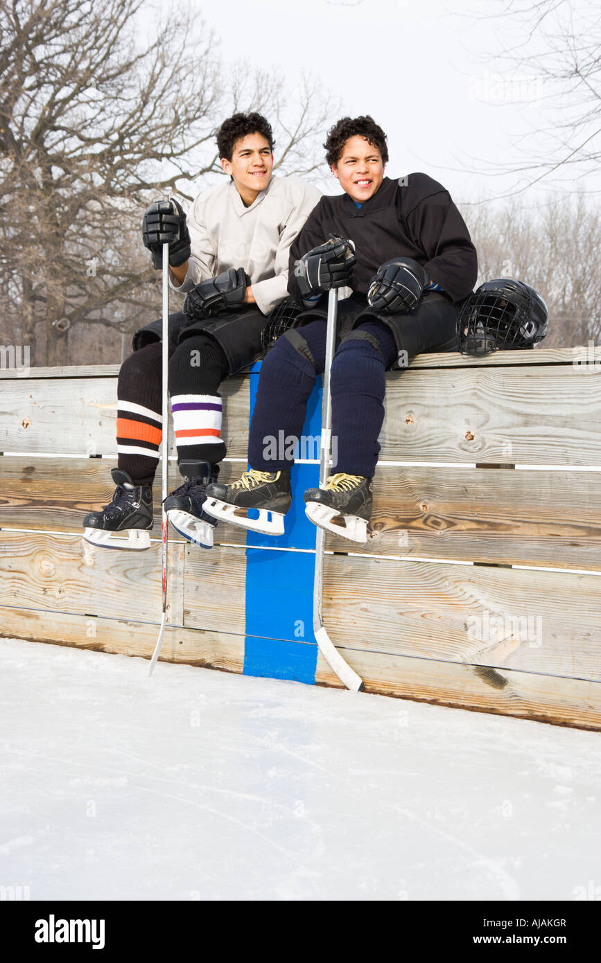 Two boys in ice hockey uniforms sitting on ice rink sidelines looking and smiling Stock Photo