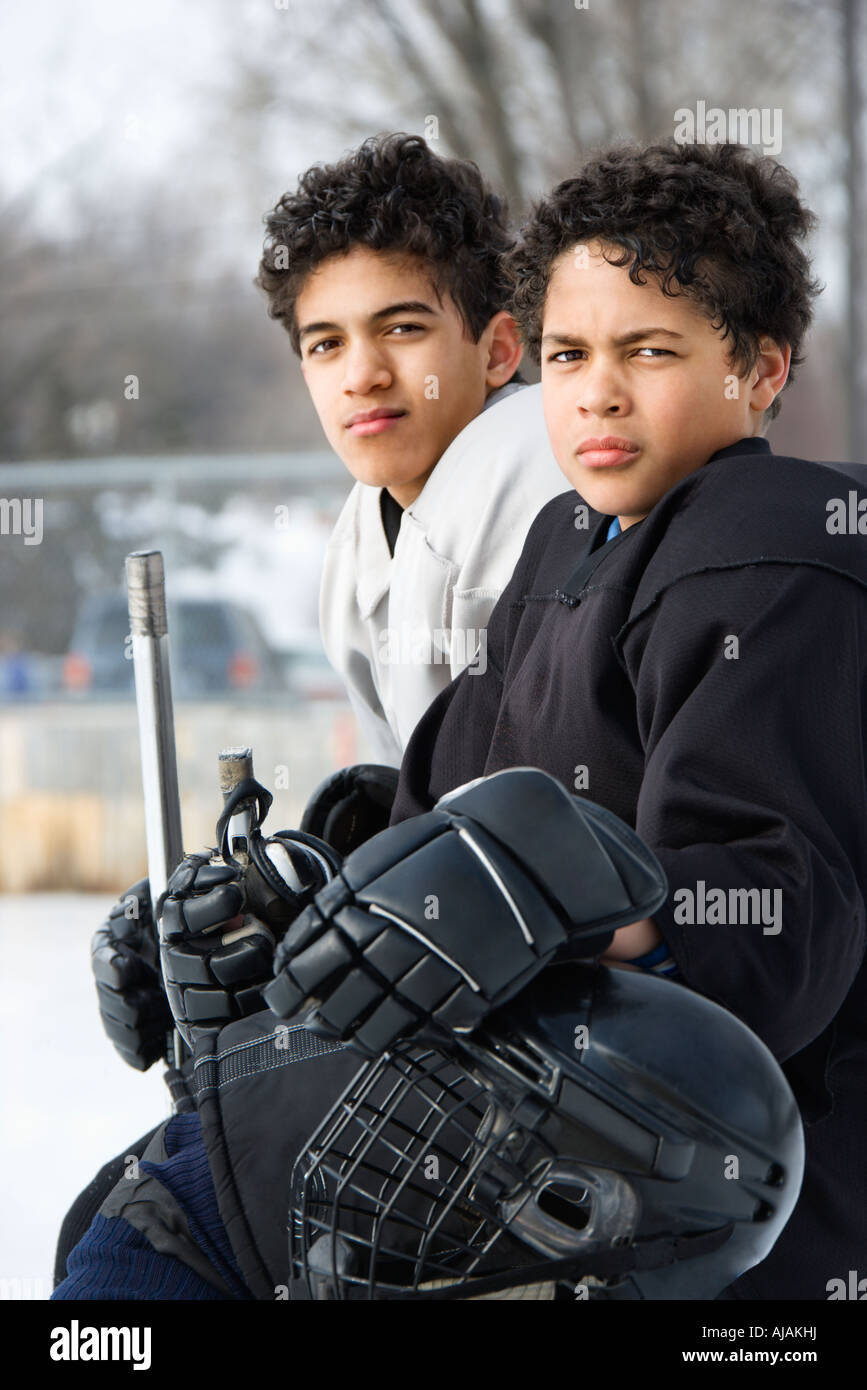 Two boys in ice hockey uniforms sitting on ice rink sidelines looking Stock Photo