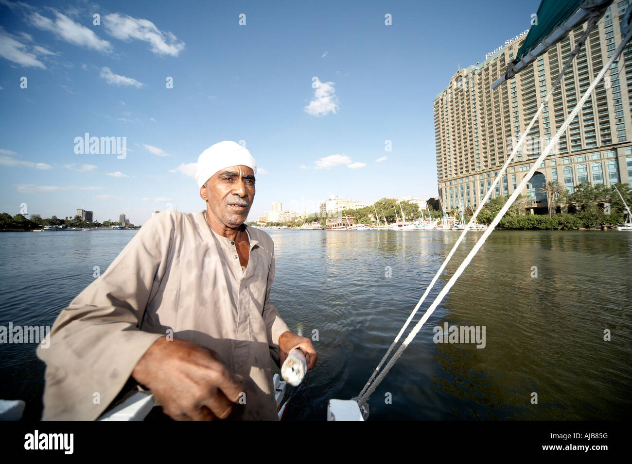 Egyptian boat man in traditional galibeya robe steering Felucca sailing boat on River Nile with Four Seasons Hotel Cairo Egypt  Stock Photo