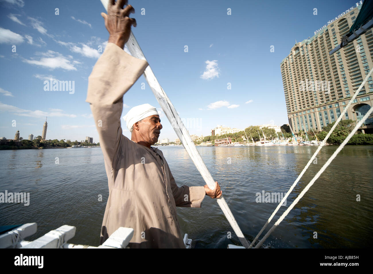 Egyptian boat man in traditional galibeya robe punting Felucca sailing boat on River Nile with Four Seasons Hotel Cairo Egypt A Stock Photo