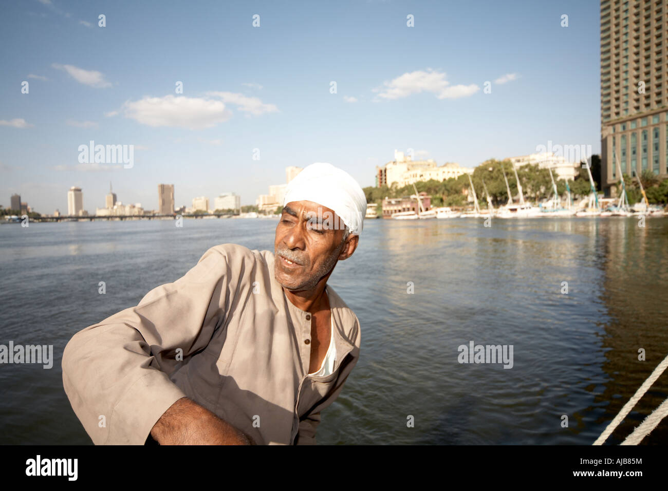 Egyptian boat man in traditional galibeya robe steering Felucca sailing boat on River Nile with Four Seasons Hotel Cairo Egypt  Stock Photo