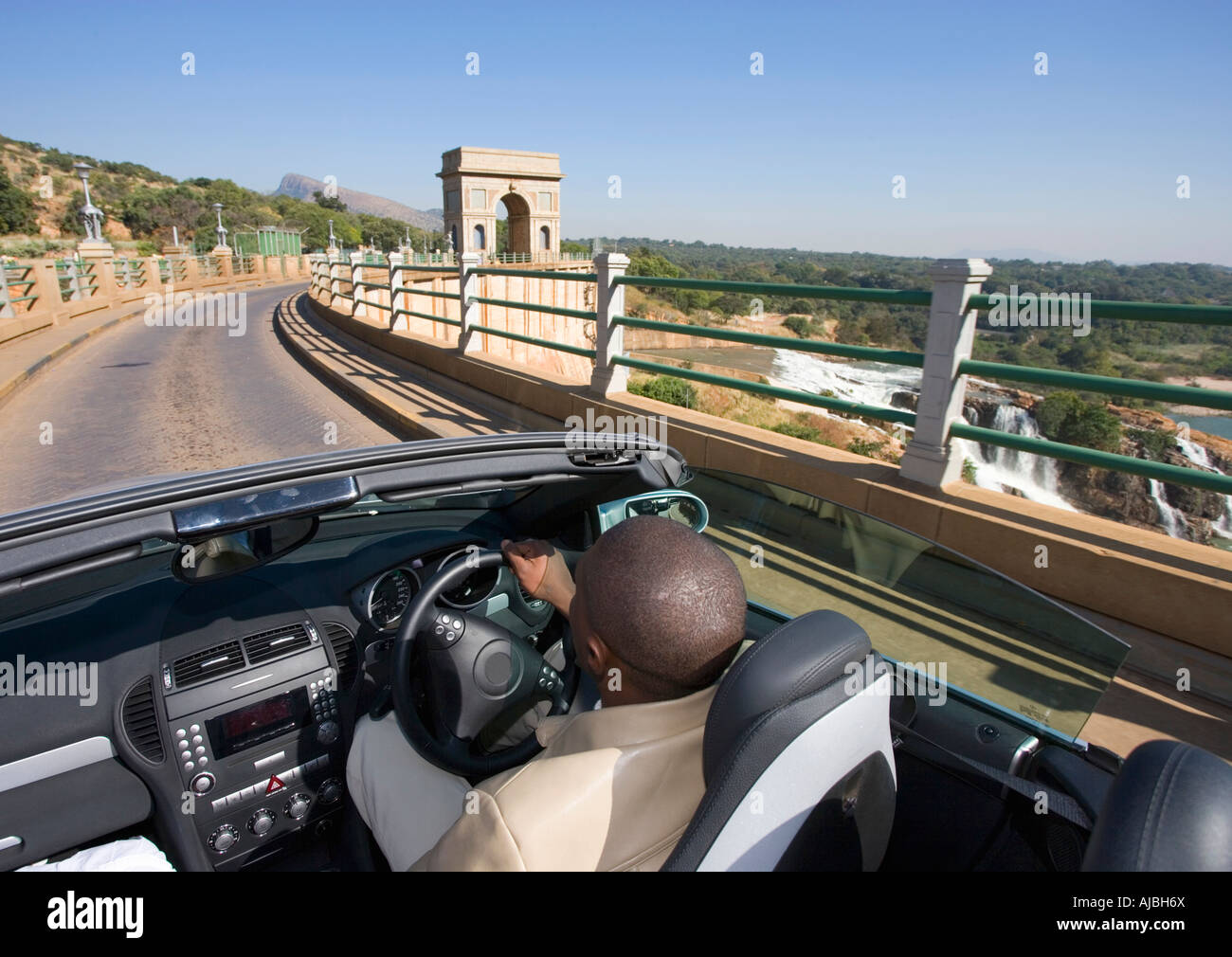 African Man Driving a Convertible Over a Dam Wall - Rear View Stock Photo