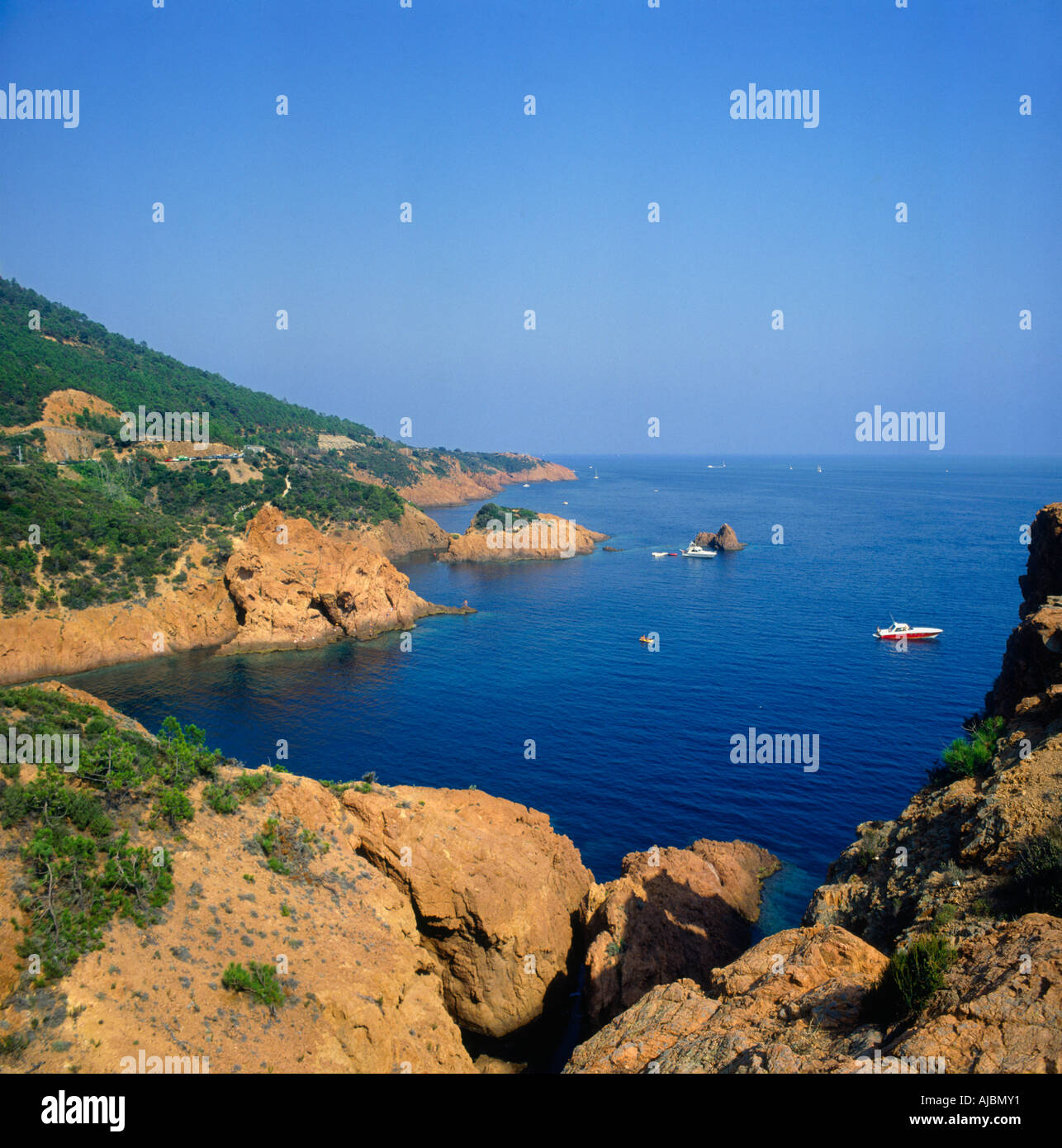 Looking across rugged rocks into blue bay with small motor launch at anchor Cap Roux Corniche de l`Esterel in South of France Stock Photo