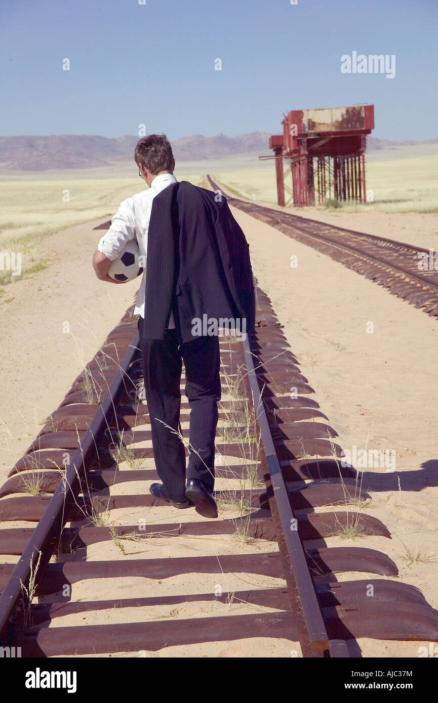 Man in a Suit Carrying a Soccer Ball & Walking Along a Desert Railroad Track Stock Photo