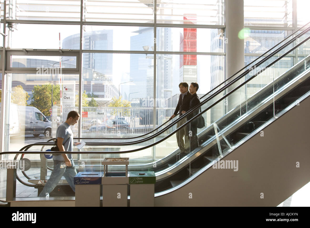 People ride an escalator in the Messe Frankfurt. Bright light enters from the glass windows behind Stock Photo
