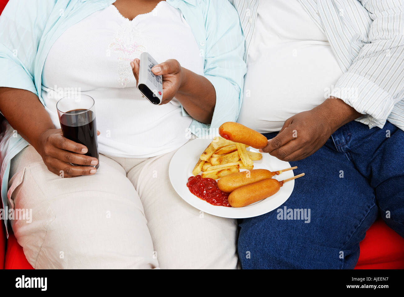 Overweight Couple, sitting, eating junk food, holding remote control, mid section Stock Photo
