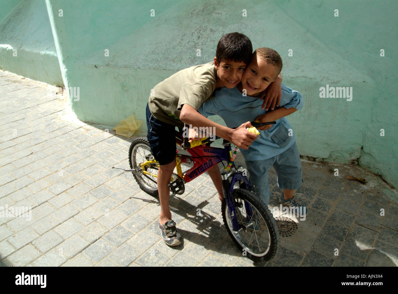 A young boy on a bike is embracing his friend and both are smiling at the camera. Stock Photo