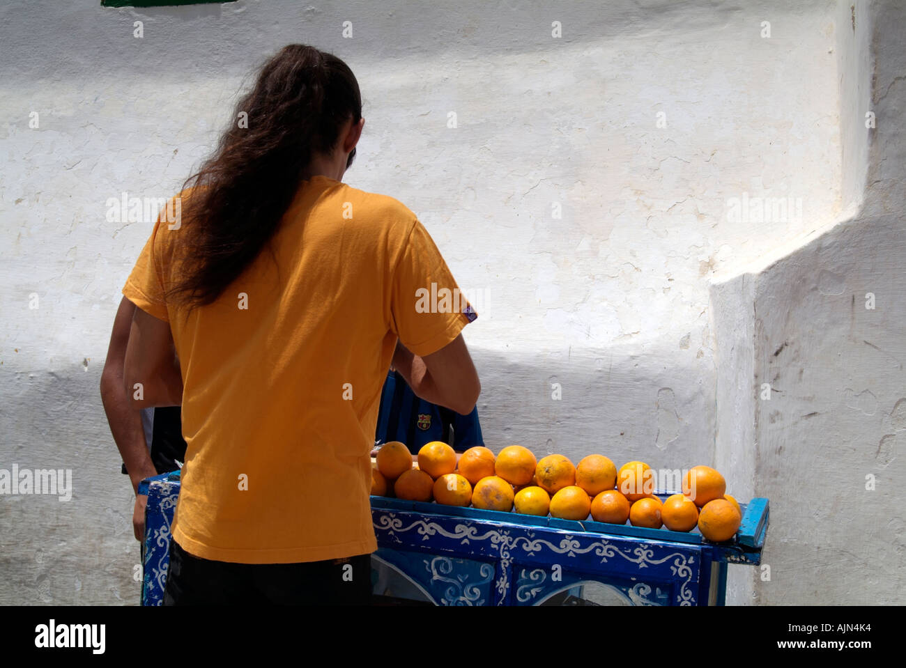 Man with his back to the camera is selling freshly squeezed orange juice from his small blue cart which is piled with oranges. Stock Photo